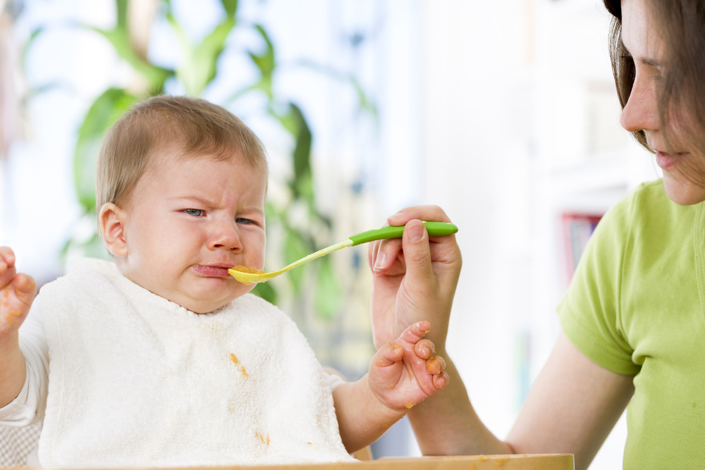 A upset baby with mother trying to feed a spoon of food to it