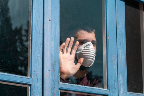 A man with his hand on the window he is looking out of wearing a mask